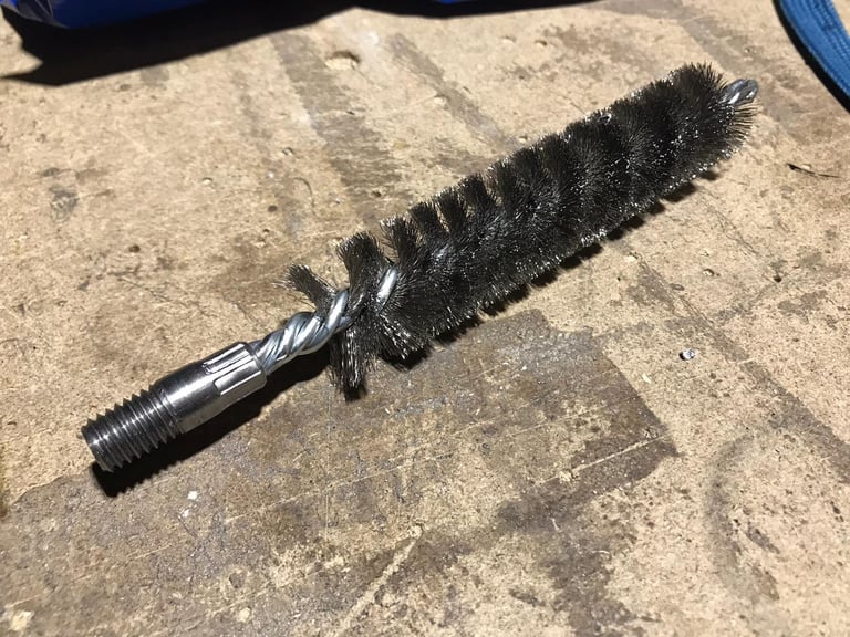 Black chimney sweep brush with bristles and threaded metal handle lying on concrete ground