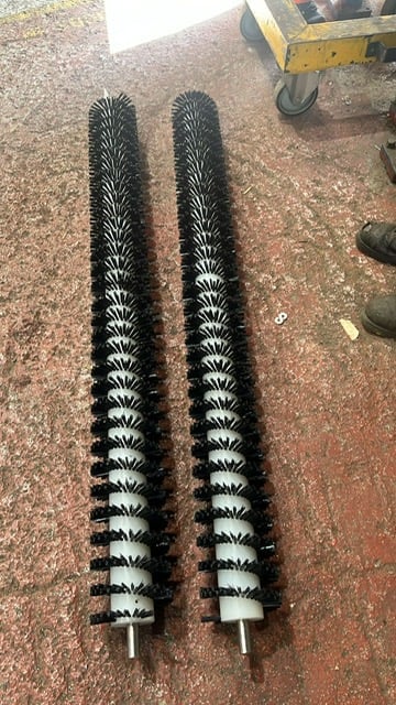 Two cylindrical street sweeper brushes with black bristles arranged parallel on a concrete surface near road maintenance equipment