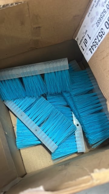 Cardboard box containing multiple blue brushes with white bristles and plastic handles, stacked for storage or shipment