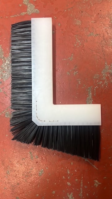 L-shaped corner broom with black bristles and white plastic handle against weathered red surface