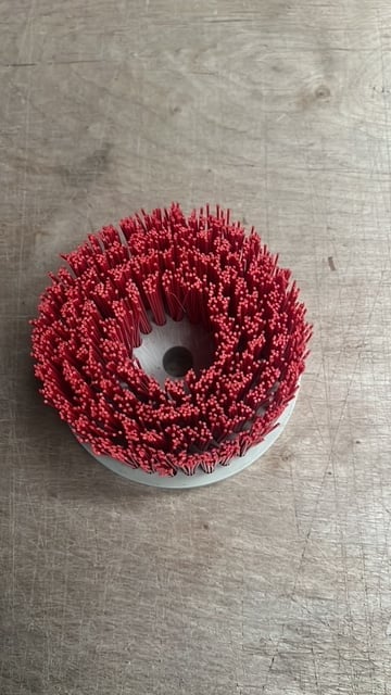Red circular abrasive brush with bristles arranged in concentric rings, mounted on white base against textured gray surface