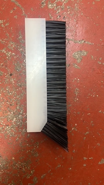 A white dustpan with black bristles leaning against a weathered red textured wall