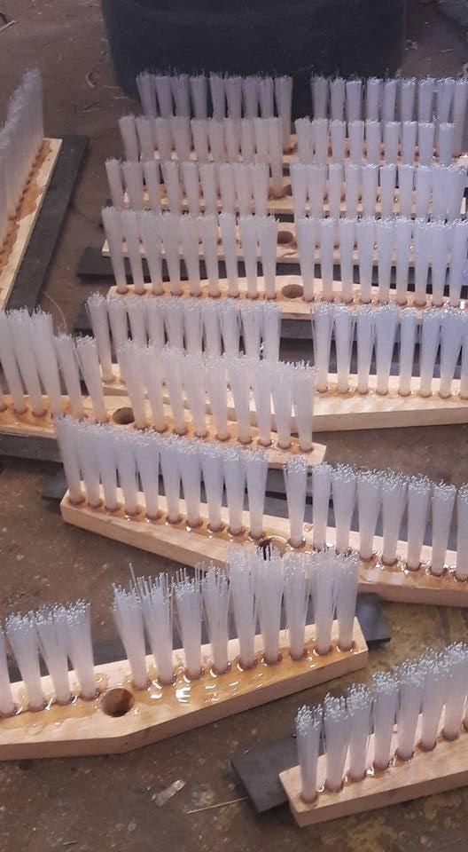 Multiple rows of white bristle brushes mounted on wooden bases in an industrial workshop or manufacturing facility