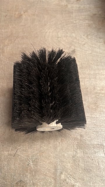 Black cylindrical brush head with stiff bristles and white plastic connector ring on beige surface
