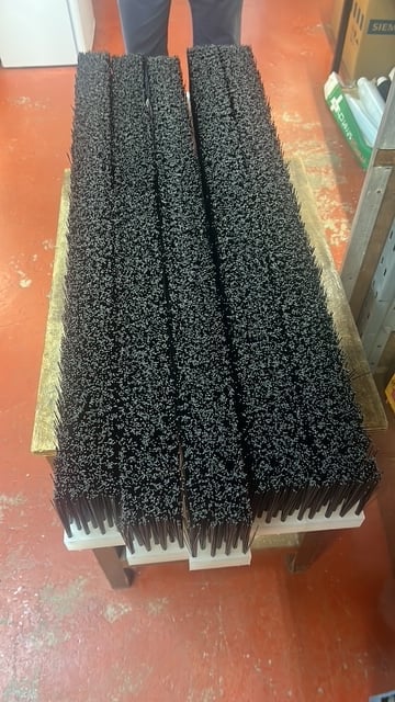 Three industrial push brooms with dark bristles and white handles stacked on an orange floor in a workshop