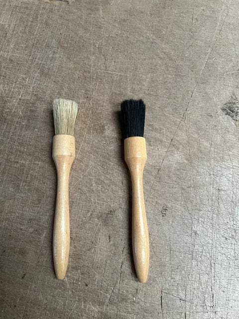 Two paintbrushes with wooden handles side by side on a textured gray surface, one with white bristles and one with black bristles