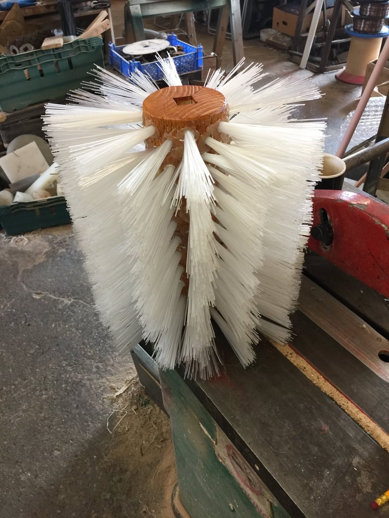 Large cylindrical brush with wooden center and white nylon bristles mounted on a work surface in an industrial workshop
