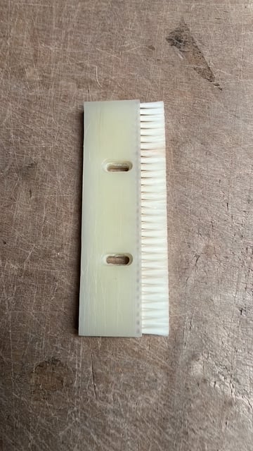 White plastic brush comb tool with two oval slots and fine bristles on textured gray surface