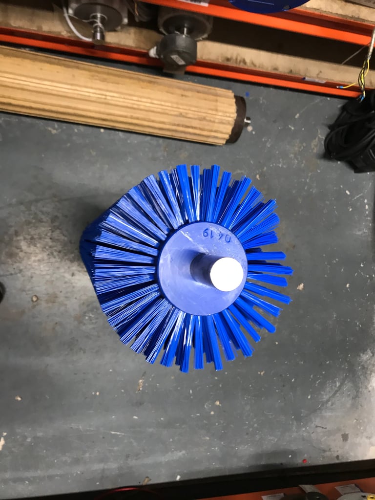 Blue circular wire brush with white center hub on dark workshop floor, near wood pole and orange shelving