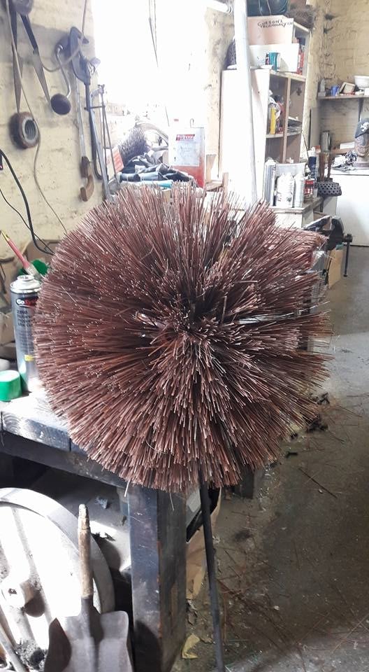 Large cylindrical wire brush with copper bristles mounted on stand in industrial workshop setting