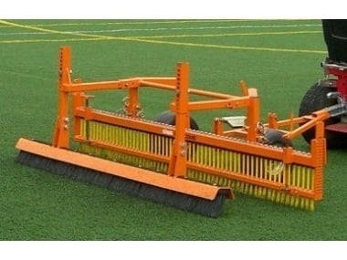 Orange farm implement with metal frame and yellow bristles attached to red tractor on green grass