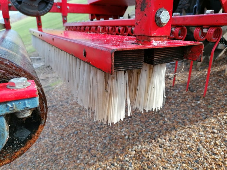 Close-up of red agricultural equipment with white bristle brushes hanging from the undercarriage, likely a field cleaning or harvesting machine