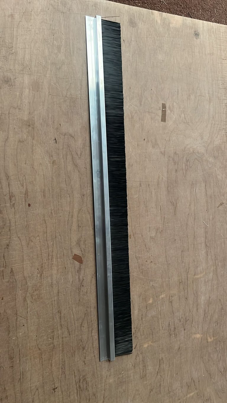 Long narrow aluminum channel with black bristle brush insert on plywood surface