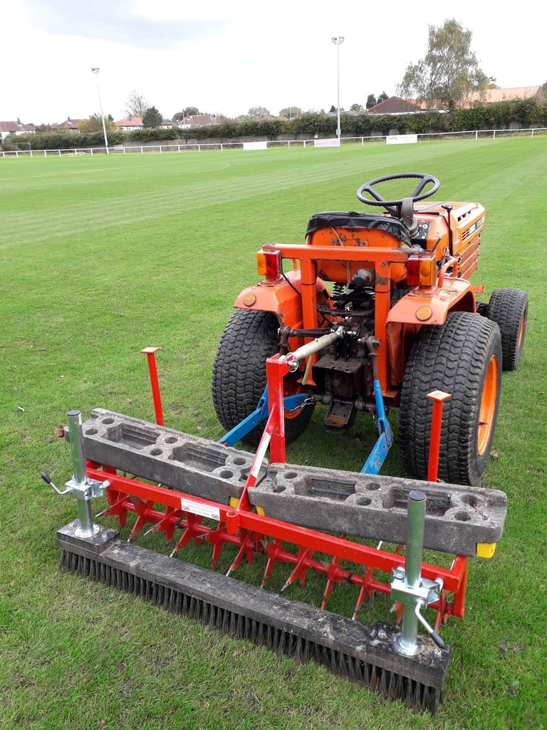 Orange ride-on mower with attached aerator and brush equipment on a sports field